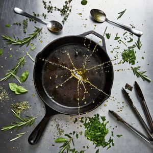 Grok Imagine output for the prompt: Overhead flat lay of a chef's station mid-service: cast iron pan, scattered herbs, a splatter of olive oil frozen mid-air