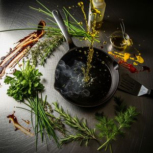 Google Image 4 output for the prompt: Overhead flat lay of a chef's station mid-service: cast iron pan, scattered herbs, a splatter of olive oil frozen mid-air