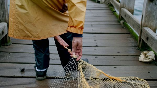 Kling 1.6 Pro output for the prompt: A elderly Japanese fisherman in a yellow raincoat mends a net on the dock. Cut to the same fisherman walking up a stone path toward a lighthouse, seen from behind. Overcast sky, 35mm film look.