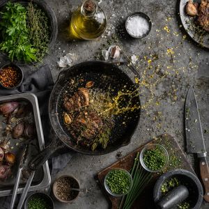 GPT Image 2 output for the prompt: Overhead flat lay of a chef's station mid-service: cast iron pan, scattered herbs, a splatter of olive oil frozen mid-air
