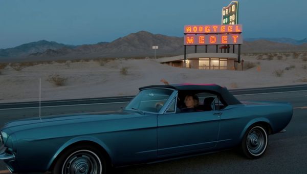 Luma Ray 2 output for the prompt: Slow orbit around a 1965 Mustang convertible parked on a Nevada desert highway at twilight. Neon motel sign blinking in the background. Cinematic widescreen.