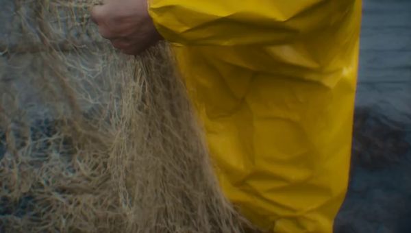 Mochi output for the prompt: A elderly Japanese fisherman in a yellow raincoat mends a net on the dock. Cut to the same fisherman walking up a stone path toward a lighthouse, seen from behind. Overcast sky, 35mm film look.