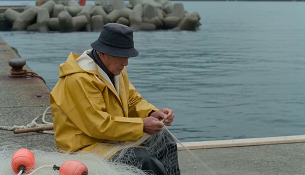 SeeDance 2.0 output for the prompt: A elderly Japanese fisherman in a yellow raincoat mends a net on the dock. Cut to the same fisherman walking up a stone path toward a lighthouse, seen from behind. Overcast sky, 35mm film look.