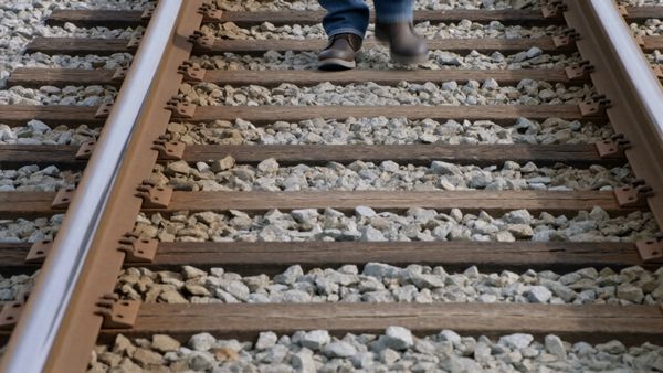 A bald man in a brown leather jacket and blue jeans is walking toward the camera along railroad tracks.  The view starts at the feet, then slowly rises to the face. The man smiles.