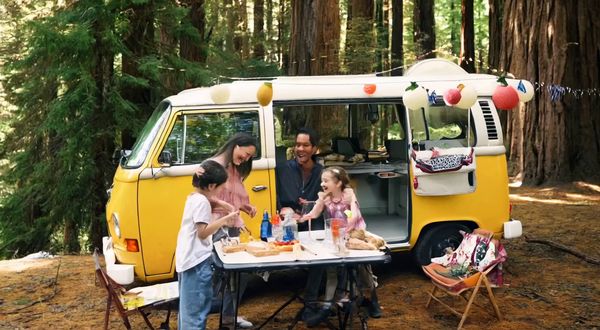 Wan v2.2 output for the prompt: Super 16mm film grain, Kodachrome color: a family of four having a picnic in a yellow VW bus parked by a redwood forest. Children laughing, mom pouring lemonade.