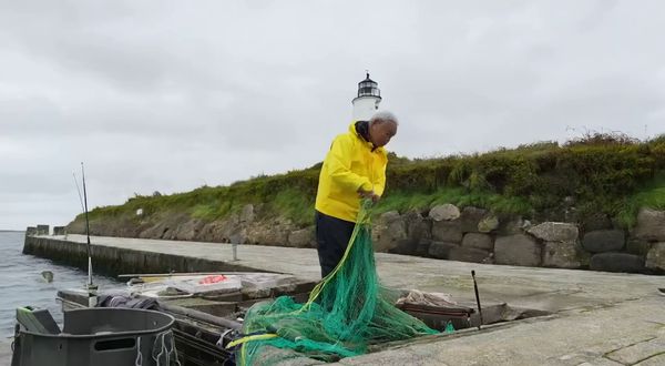 Wan v2.2 output for the prompt: A elderly Japanese fisherman in a yellow raincoat mends a net on the dock. Cut to the same fisherman walking up a stone path toward a lighthouse, seen from behind. Overcast sky, 35mm film look.