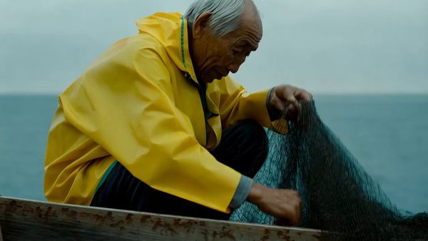Minimax output for the prompt: A elderly Japanese fisherman in a yellow raincoat mends a net on the dock. Cut to the same fisherman walking up a stone path toward a lighthouse, seen from behind. Overcast sky, 35mm film look.