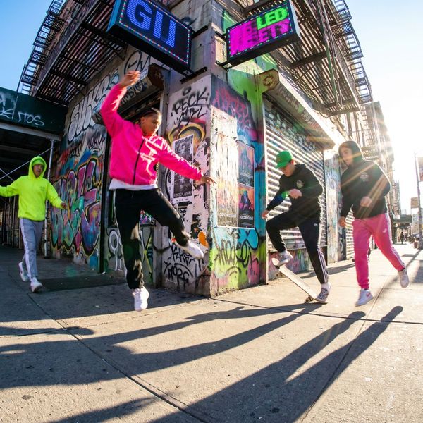 High-energy wide-angle street photography style with extreme fisheye distortion shot from low angle, featuring direct harsh sunlight creating sharp shadows and saturated neon colors—electric blues, hot pinks, lime greens—with intense contrast. Figures in spontaneous motion with slight blur against concrete and graffitied urban surfaces. Candid youth culture and street fashion moments captured raw and visceral, emphasizing dynamic energy and attitude. Generate random, unpredictable scenes each time.